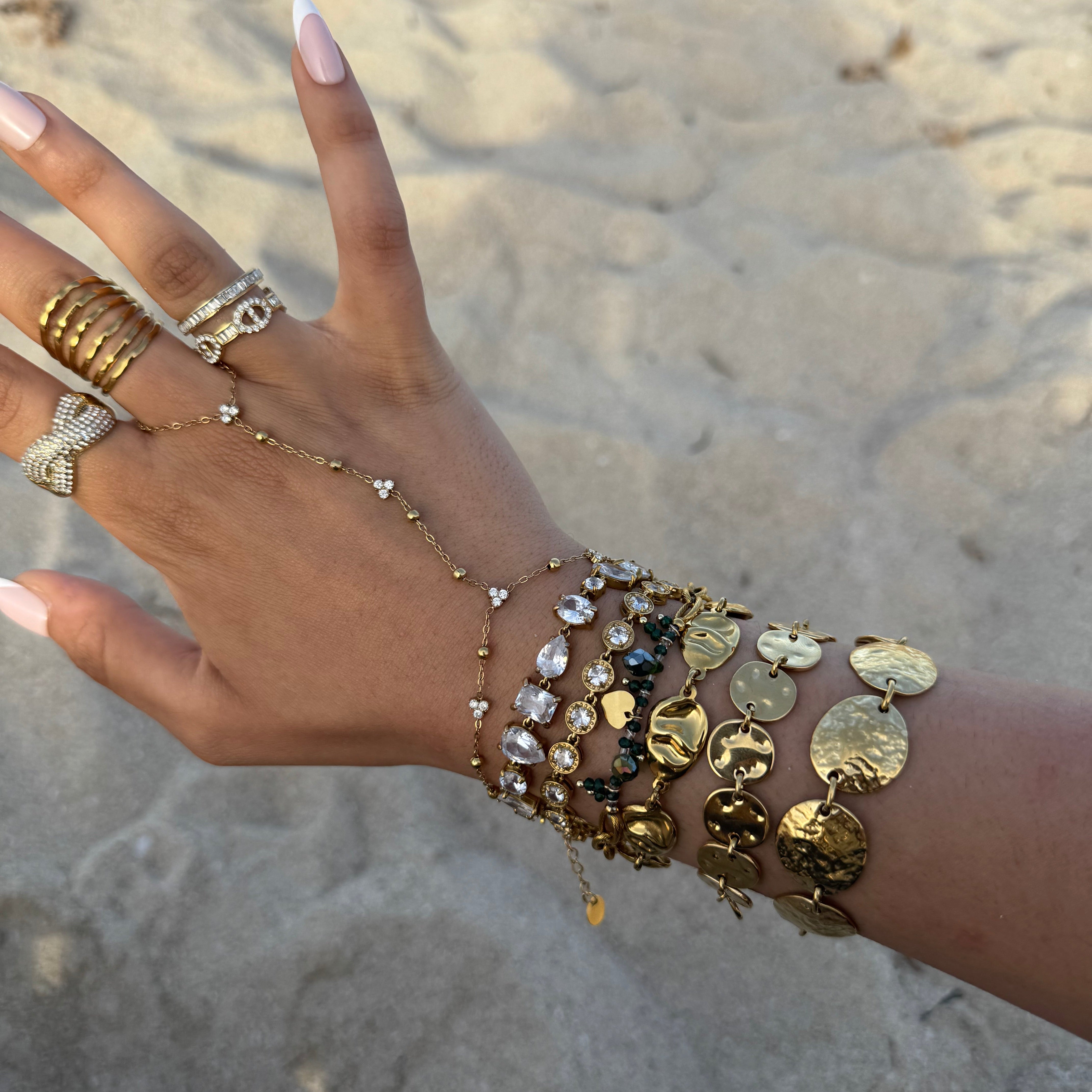 Gros plan d'une main délicate ornée d'une chaîne de main dorée et de multiples bracelets médailles sur une plage de sable clair.