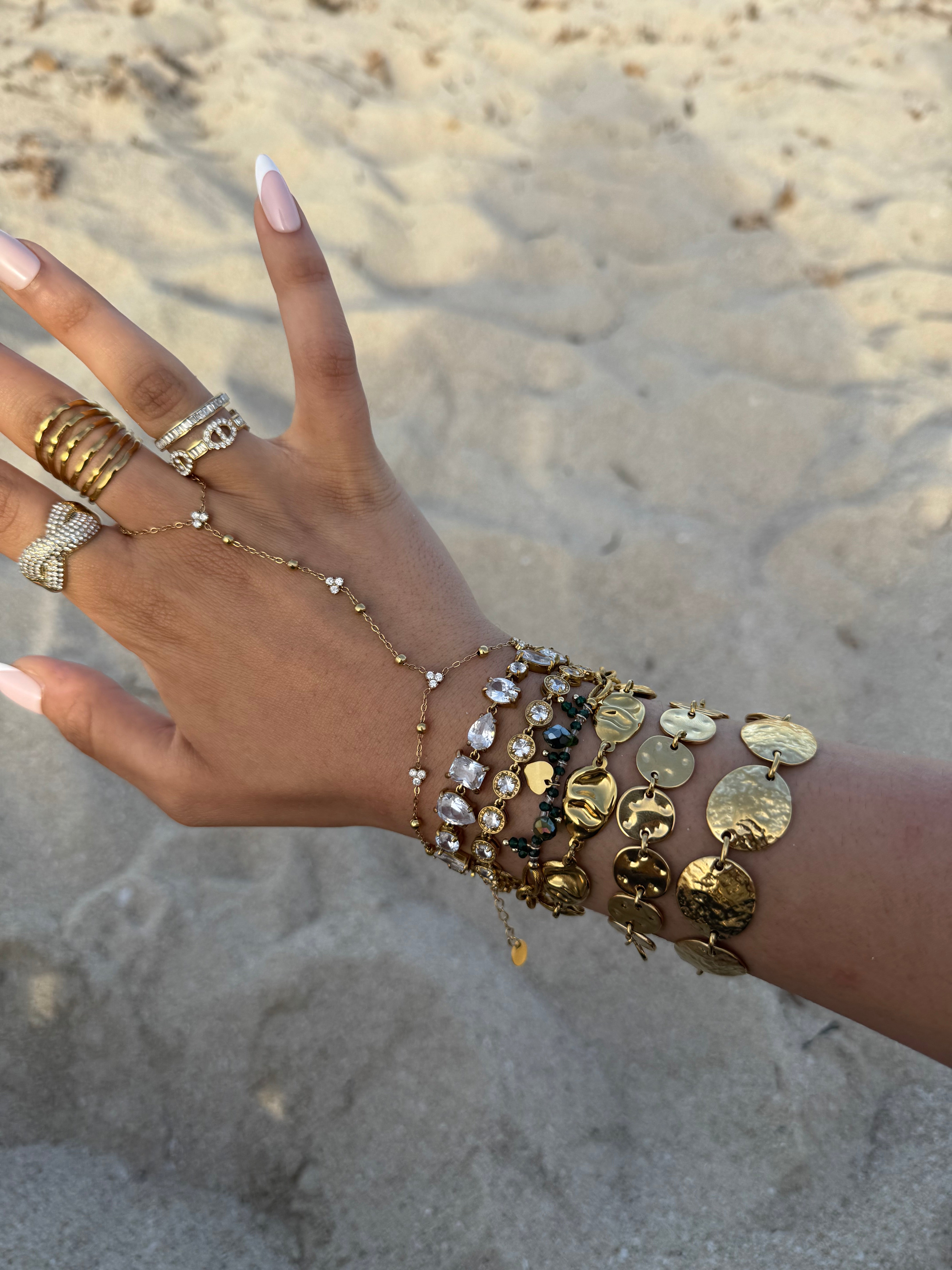 Gros plan d'une main délicate ornée d'une chaîne de main dorée et de multiples bracelets médailles sur une plage de sable clair.