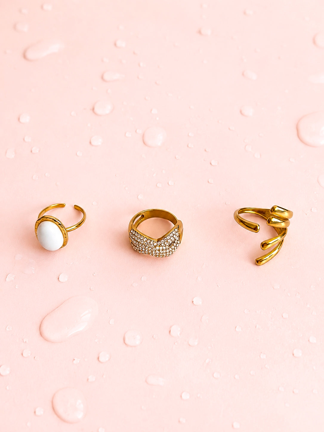 Trois bagues dorées en acier inoxydable pour femme avec pierre blanche, strass et design torsadé sur fond rose goutte d'eau