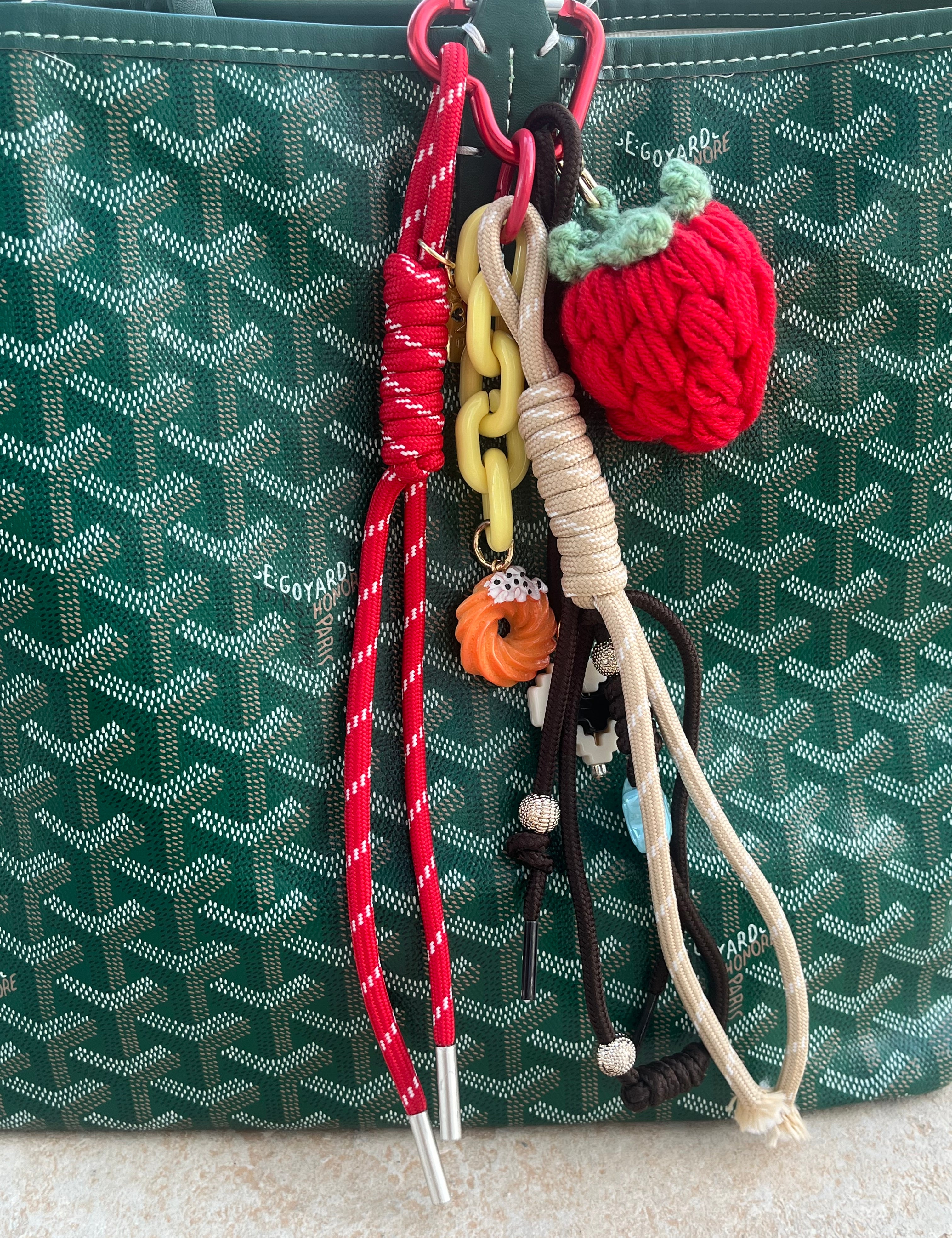 Grigri Velvet Sunset à fraise en crochet et cordons rouge et beige, accroché à un sac vert - Ordiajade