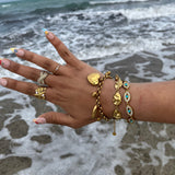 Bague Rym ajustable portée à la main avec d'autres bijoux dorés en bord de mer.