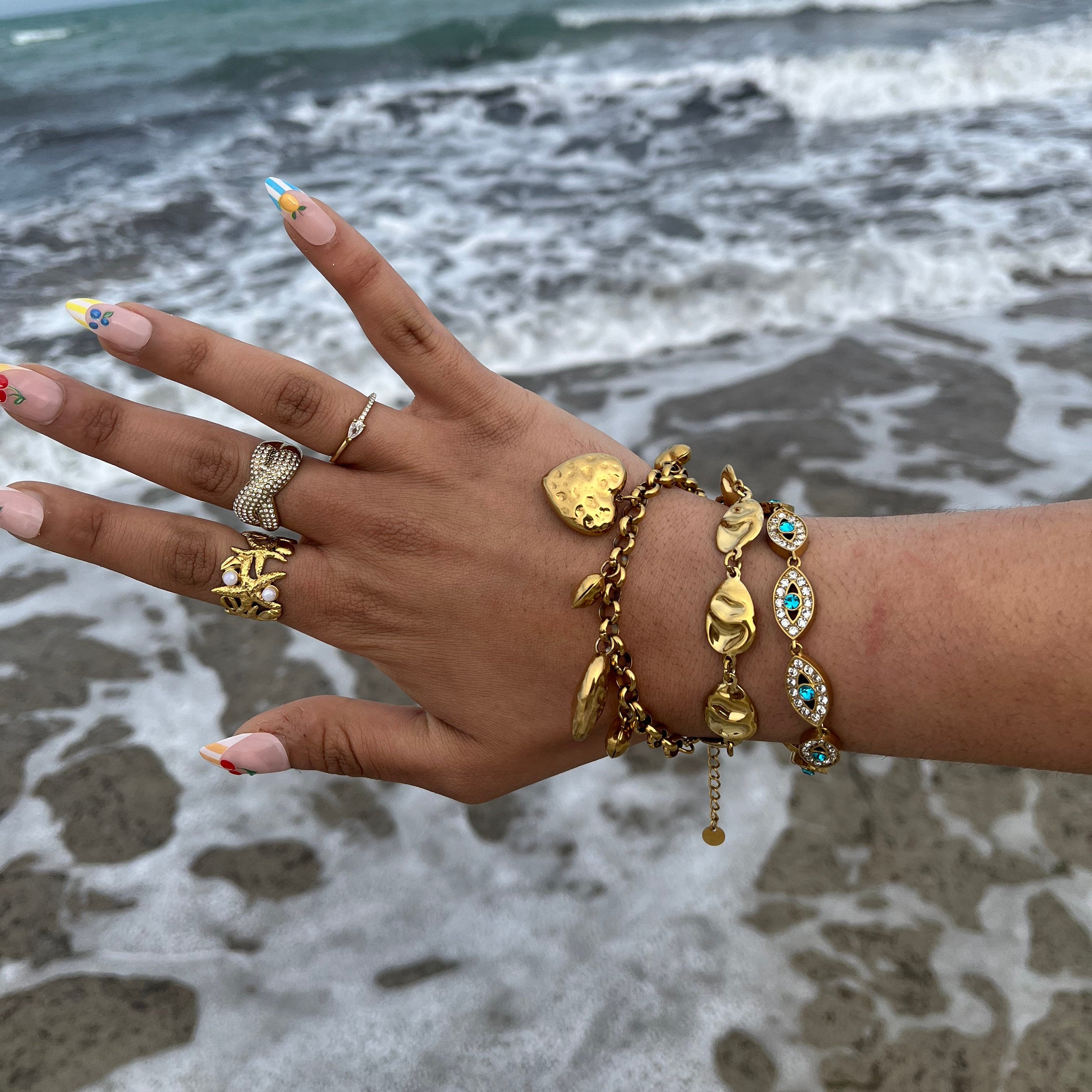 Bague Rym ajustable portée à la main avec d'autres bijoux dorés en bord de mer.