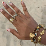 Bague Rym ajustable portée à la main avec d'autres bijoux dorés en bord de mer sable