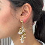 Grandes boucles d'oreilles Rich dorées avec motifs fleurs blanches et perles nacrées portées de profil.