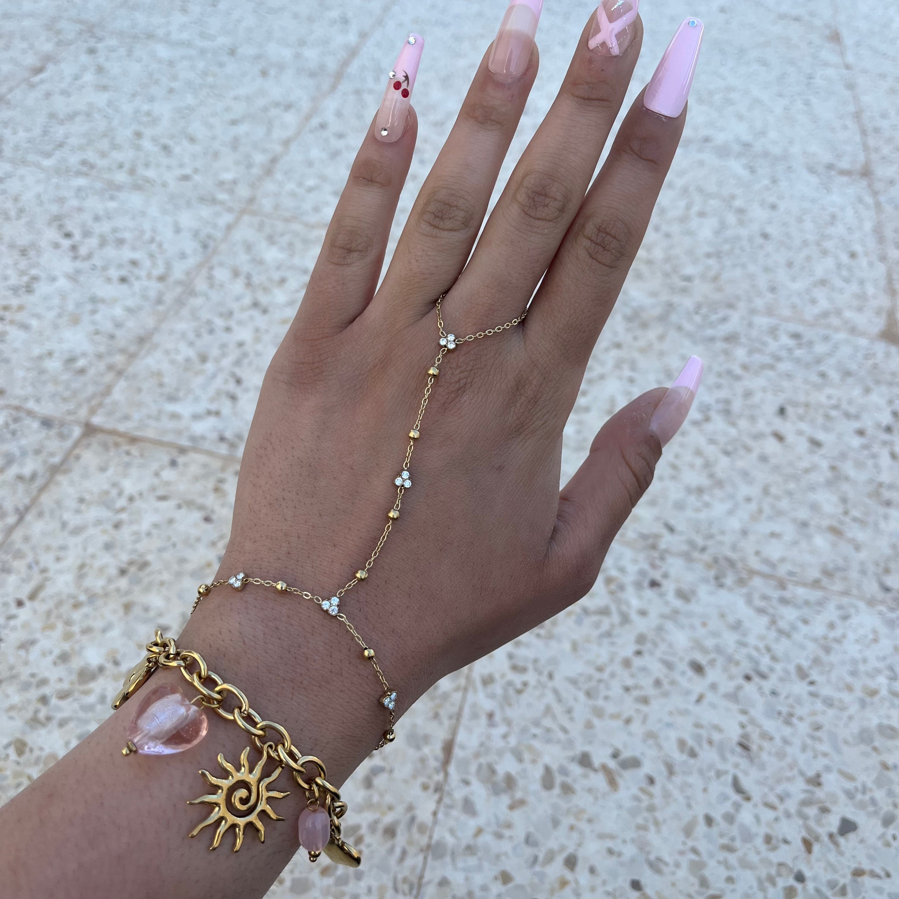 Photo portée de la chaîne de main Aiko dorée avec de petites perles et des zircons, présentée sur une main avec une manucure rose.