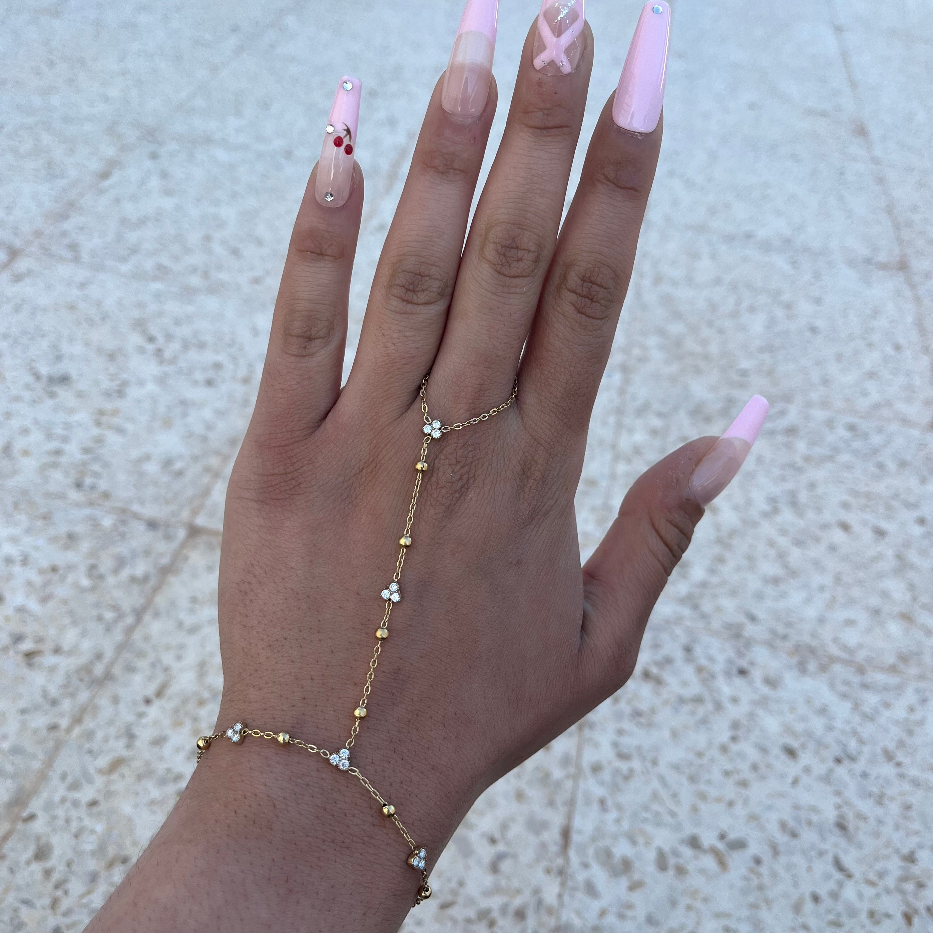 Photo portée de la chaîne de main Aiko dorée avec de petites perles et des zircons, présentée sur une main avec une manucure rose.